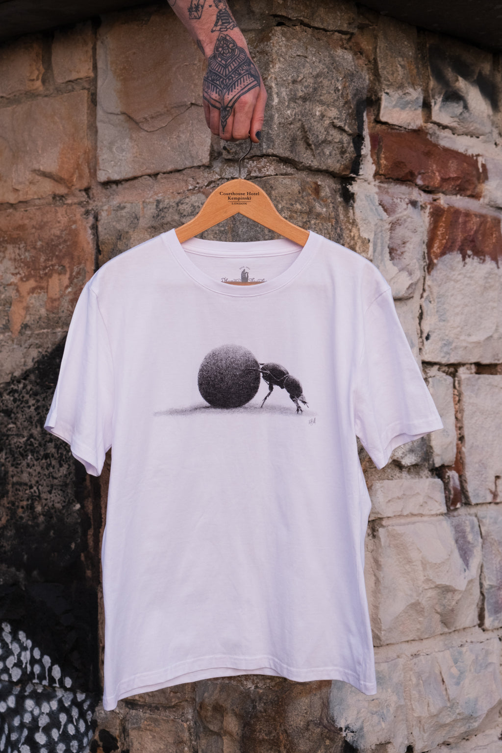 White t-shirt with a graphic design of a dung beetle pushing a ball hanging on a wooden hanger against a stone wall.